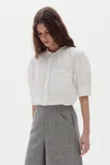 Camisa blanca de algodón con cuello mao, botonera oculta, mangas cortas estilo raglán con cortes estructurados y un bolsillo frontal con cierre.
