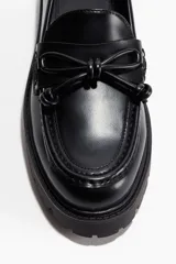 Mocasines negros de suela gruesa con costura frontal y detalle de tiras decorativas cruzadas con nudos.