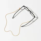 Correa para lentes dorada con cadena de eslabones trenzados y terminales de goma transparente ajustables. Incluye dije circular con el nombre de la marca grabado.