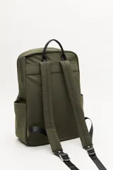 Mochila urbana de nylon en color verde militar, con detalles en cuero sintético negro. Cuenta con compartimento principal con cierre, bolsillo frontal con cierre, bolsillos laterales abiertos y asa superior. Incluye un colgante decorativo de tela y correas ajustables acolchadas.