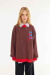 Sweater de punto color amarillo con silueta oversize y estilo universitario vintage. Presenta cuello redondo, puños y ruedo en tejido acanalado de color gris azulado, y un diseño gráfico bordado en el frente.