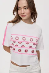 Remera blanca de manga corta, con cuello redondo, calce relajado y estampado frontal con motivos gráficos de soles y símbolos marinos en tonos rosados y bordo, además de líneas horizontales.