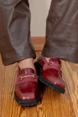 Mocasín de cuero vacuno color marrón, con suela track de goma negra, puntera cuadrada y detalle de herraje metálico sobre una banda de gamuza en el empeine.