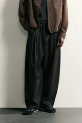 Pantalón de corte balloon fit en color gris oscuro, confeccionado en tejido con viscosa. Presenta pliegues frontales, cintura ajustable con cinturón integrado, bolsillos laterales y bolsillos traseros de doble vivo. Cierre frontal con cremallera y botón.