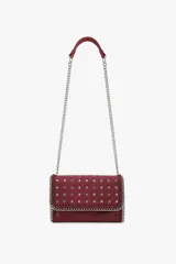 Cartera tipo bandolera de gamuza color rojo oscuro, con diseño rectangular y solapa frontal decorada con tachas metálicas en forma de estrella. Presenta bordes reforzados con cadena metálica plateada y correa larga de cadena con hombrera de gamuza a tono.