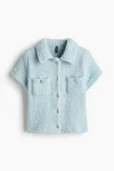 Saco corto de manga corta color celeste confeccionado en sherpa suave. Presenta cuello camisero, cierre frontal de botones metálicos dorados, bolsillos de parche en el pecho con solapa y botones, y diseño de bajo recto.