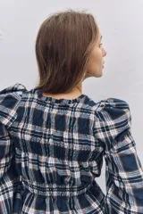 Blusa de manga larga con diseño a cuadros escoceses en tonos azul marino, blanco y naranja. Presenta cuello redondo, hombros ligeramente abullonados y detalle de fruncido elástico en la cintura que crea un efecto peplum.
