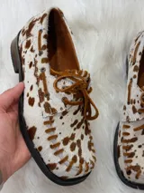 Zapato tipo mocasín de cuero con pelo estampado con diseño de manchas de vaca en tonos marrón y blanco. Presenta cordones frontales, costuras visibles en el borde y suela plana.