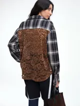 Camisa de corte relajado con estampado de cuadros en tonos negro y beige. Presenta cuello camisero, cierre frontal parcial con botones y mangas largas con puños ajustables mediante lazos. El diseño destaca por un detalle de encaje en la espalda que aporta contraste.