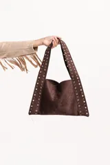 Cartera tipo tote de gamuza color tostado, con diseño triangular y correas integradas decoradas con tachas metálicas doradas a lo largo de los bordes.
