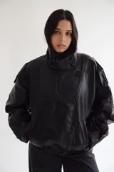 Chaqueta negra de efecto cuero, estilo bomber o motociclista, con cuello alto abotonado y cierre frontal asimétrico. Presenta detalles de costuras verticales y hombreras pequeñas. El bajo es elástico.