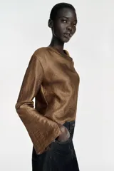 Blusa de manga larga con cuello fluido tipo drapeado y abertura en los puños. Presenta un acabado satinado con textura y cierre posterior con cremallera oculta.