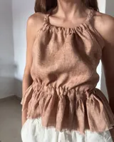 Blusa color salmón/rosado claro, confeccionada en lino, con escote recogido y tiras trenzadas regulables que cruzan en la espalda. Presenta un elástico fruncido en la cintura que da lugar a un pequeño volante con terminación deshilachada.