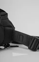 Bolso cruzado (bandolera) negro de tela sintética, con correa ajustable estampada con la palabra "LUXO". Presenta un diseño estructurado con bolsillo frontal y parche con logo en la base.