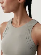Top deportivo color gris con perforaciones decorativas en la parte delantera y trasera. Tejido Shape_Tech con tecnología performance, de sujeción media-alta, con copas extraíbles.