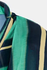 Camisa de manga larga con escote fluido tipo pañuelo, hombreras y puños abotonados. Presenta un diseño de rayas verticales en tonos verdes y amarillos.