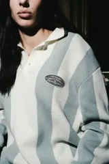 Chomba de manga larga con diseño de rayas verticales en color blanco y verde seco. Presenta cuello tipo polo con cierre de botones y parches decorativos en el pecho.