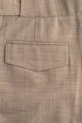 Pantalón sastrero de corte amplio y tiro alto, color beige/tostado con textura sarga. Presenta pinzas frontales y pliegues marcados en las piernas. Tiene cierre con botón y corchete ocultos, y detalles de ajuste con botones forrados en la cintura trasera.
