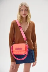Bandolera acolchada color coral con solapa y cierre magnético. Tiene correa ancha de tela rosa con logo de la marca y asa de mano trenzada en color rosa.