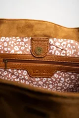 Bolso tipo tote de gran tamaño confeccionado en gamuza sintética color tostado. Presenta un diseño minimalista con asas largas de hombro y una pequeña placa metálica frontal con el logo de la marca.
