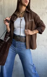 Camisa de gamuza color chocolate con bordados decorativos en color crudo a lo largo de la botonadura frontal y en el bolsillo superior. Presenta cuello clásico, cierre frontal con botones y mangas largas.