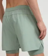 Bermuda deportiva para hombre, color verde menta, con short de compresión integrado. La bermuda exterior es suelta y tiene un pequeño logo estampado en el muslo izquierdo.