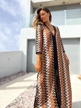 Vestido largo de tejido de punto con diseño geométrico en tonos tierra, negro y blanco. Presenta escote en V profundo, mangas tres cuartos y tajos laterales en la falda.