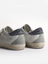 Zapatillas Golden Goose modelo Super-Star de cuero blanco con estrella de gamuza gris, refuerzo de talón con purpurina gris oscuro, aplicación trasera de gamuza gris y cordones color crema.