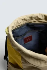 Mochila confeccionada en tejido técnico con bloques de color beige, amarillo y verde, y detalles en piel. Cuenta con un compartimento principal con cierre de solapa, hebilla y cordón ajustable, bolsillos laterales planos y correas de hombro regulables con hebilla de liberación rápida.