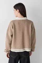 Cardigan de lana color crema con cuello redondo, cierre frontal de botones y mangas largas con puños ajustados. Presenta bordados decorativos en el frente que incluyen un sol, olas y estrellas, con detalles en azul en el cuello y el ruedo.