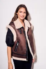 Chaleco oversize estilo gamulán en color marrón, confeccionado en cuero vegano con forro y solapas de corderito sintético en contraste. Posee cierre frontal metálico, bolsillos con cremallera y detalles de correas con hebillas en el cuello.