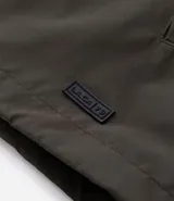 Campera rompevientos color tostado, con capucha ajustable mediante cordones, cierre frontal completo y puños elásticos. Presenta un diseño minimalista y funcional, ideal para protección contra el viento.