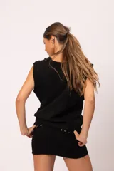 Pollera mini de denim negro con terminación desflecada. Presenta un diseño decorado con múltiples tachas cuadradas plateadas de diversos tamaños distribuidas en la cintura, sobre las solapas de los bolsillos delanteros y en el frente.