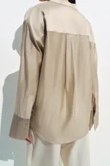 Camisa de cuello mao color beige, confeccionada en tejido satinado con brillo sutil, manga larga con puños anchos y un bolsillo de parche en el pecho con detalles bordados tonales. Cierre frontal con botones ocultos bajo solapa.