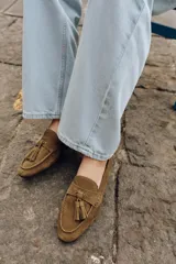 Mocasín de mujer confeccionado en serraje color marrón, con detalle de antifaz y borlas en el empeine. Presenta punta redonda y suela plana.