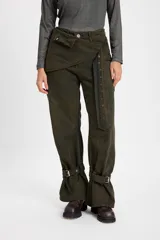 Jean de corte baggy con pierna recta y ancha, color verde oliva. Presenta un diseño de cintura cruzada con solapa y tira decorativa con botones metálicos. Incluye tiras ajustables con hebillas en los tobillos, costuras reforzadas y bolsillos funcionales.