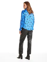 Camisa de satén de corte holgado con cuello camisero y cierre frontal de botones metálicos. Presenta un estampado de cuadrados negros sobre fondo azul vibrante, mangas largas con puños abotonados y un acabado brillante característico del satén.