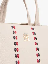 Cartera tipo satchel de Tommy Hilfiger en color beige, confeccionada en poliuretano con textura granulada. Presenta doble asa superior, correa ajustable extraíble, logo TH metalizado en el frente y dos franjas verticales decorativas con detalles entrelazados en rojo y negro.