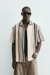 Camisa de manga corta con calce relaxed fit, confeccionada en tejido estructurado con algodón. Presenta un diseño de rayas verticales en tonos beige, negro y blanco, cuello solapa, cierre frontal con botones y aberturas laterales en el bajo.
