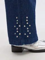 Pantalón jean de pierna ancha, color azul oscuro, con pinzas y detalles de tachas en los bajos. Presenta bolsillos laterales y traseros tipo sastrero.