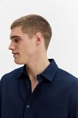 Camisa rústica de manga larga color gris oscuro, con cuello clásico, cierre frontal con botones y un bolsillo aplicado en el pecho.