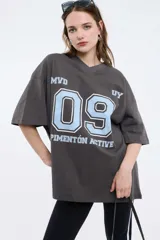 Remera oversize gris oscuro con cuello en V y mangas cortas. Presenta una estampa frontal estilo universitario con el número "09" y las inscripciones "MVD", "UY" y "PIMENTÓN ACTIVE" en tonos celestes y blancos.