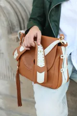 Cartera tipo bandolera de cuero en color camel con detalles en cuero blanco. Presenta una tapa frontal con apliques de tachas metálicas doradas, correas laterales decorativas y un detalle de flecos largos en uno de los laterales. Incluye herrajes dorados y correa ajustable.