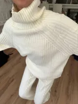 Polerón blanco de cuello alto, tejido de punto con textura acanalada y detalles de trenzas en las mangas.