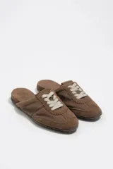 Zapatilla estilo mula de color beige, confeccionada en una combinación de algodón y serraje vacuno, con suela de caucho con tachuelas texturizadas y costuras decorativas visibles en la parte superior.