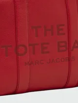 Bolso tipo tote de cuero negro, modelo The Leather Medium Tote Bag de Marc Jacobs. Presenta doble asa superior, correa ajustable para el hombro y el texto grabado "THE TOTE BAG MARC JACOBS" en el frente.