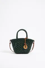 Bolso tipo cesta de mano, confeccionado en material sintético trenzado de color verde oscuro. Presenta asas rígidas redondeadas, una correa larga ajustable de color marrón y un dije circular decorativo con logo grabado.