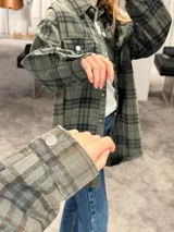 Camisa tipo sobrecamisa de corte oversize, confeccionada en tela a cuadros en tonos marrones y negros. Presenta cierre frontal con botones metálicos a presión, bolsillos frontales con solapa y mangas largas con puños abotonados.