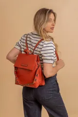 Mochila tipo cartera con tapa, confeccionada en símil cuero color suela. Cuenta con doble compartimento, división interna, cierre de seguridad y bolsillo trasero con cierre. Incluye una correa frontal decorativa con tachas metálicas.