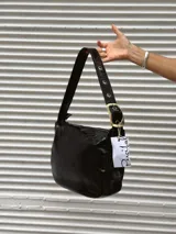 Bolso de hombro tipo hobo en color negro con acabado brillante. Presenta una correa ajustable con ojales metálicos y una hebilla decorativa grande en tono dorado. Cuenta con cierre superior y un diseño práctico para uso diario.